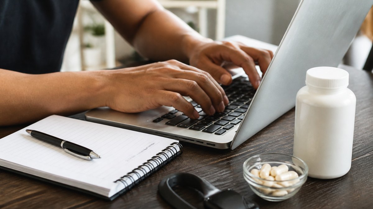 Close-up of a person's hands typing on a laptop at a wooden desk, with a notepad, pen, and a bottle of dietary supplements beside the keyboard, suggesting someone researching manufacturer records online