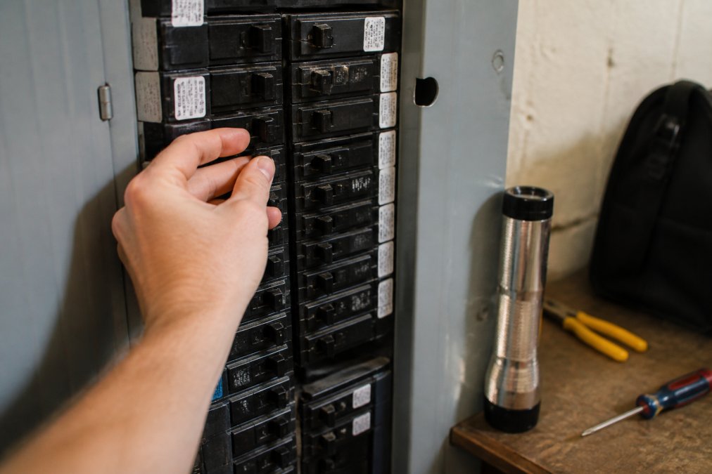 Close-up of a homeowner's hand flipping a breaker switch inside an open electrical panel, with labeled breakers visible and a flashlight nearby