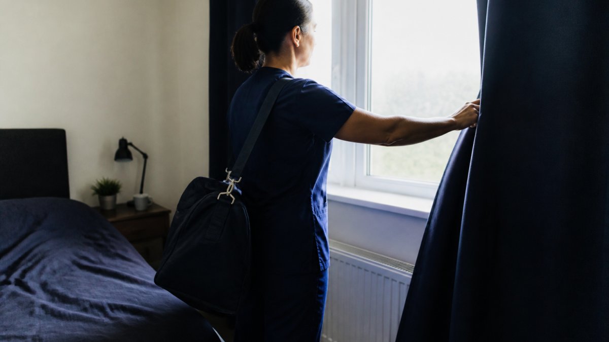 A person arriving home after a night shift, closing thick bedroom curtains to block out bright morning daylight before sleeping