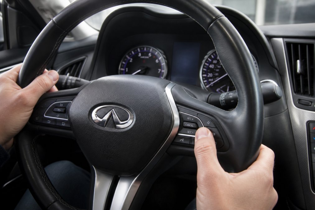 Close-up of a driver's hands using steering wheel buttons to navigate the dashboard display in an Infiniti Q50, with the instrument cluster visible and a maintenance menu on screen
