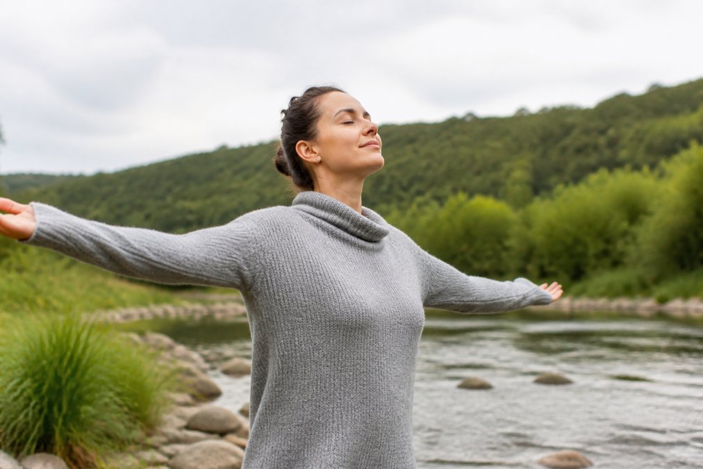 Person standing outdoors in a calm, natural setting with arms gently extended, demonstrating a somatic release posture with relaxed shoulders and open body language