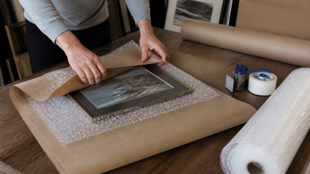 Hands carefully wrapping a framed artwork in bubble wrap and paper on a workspace with packing materials