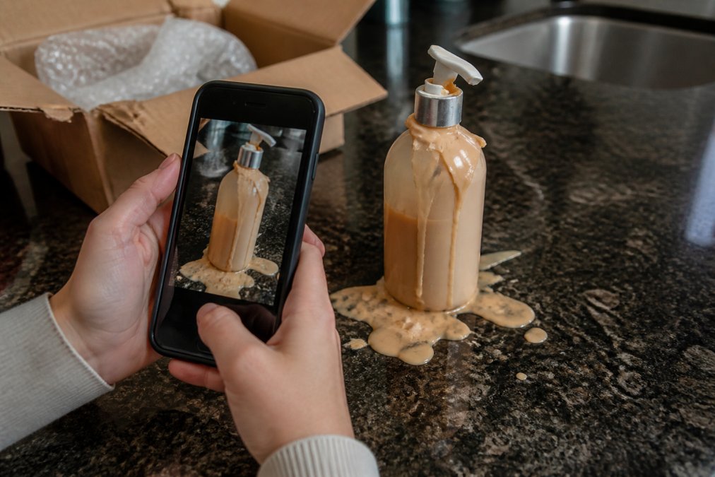Close-up of hands holding a smartphone taking a photo of a damaged, leaking cosmetic bottle with a shattered pump, placed on a kitchen counter with the shipping box visible in the background