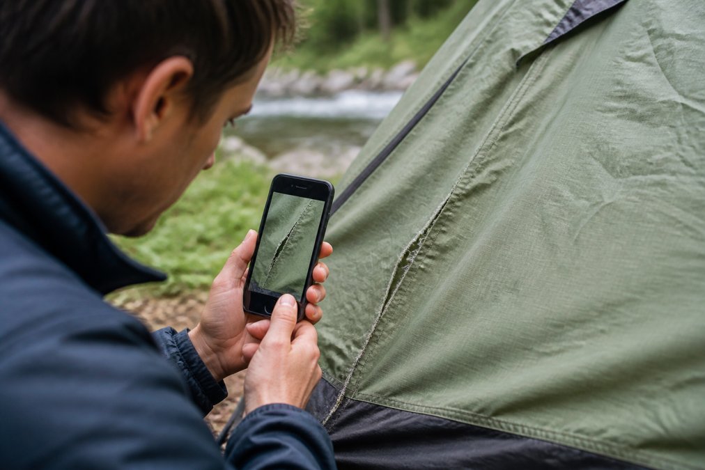 A person photographing a ripped tent seam with a smartphone, focusing on the damage to document for a return claim