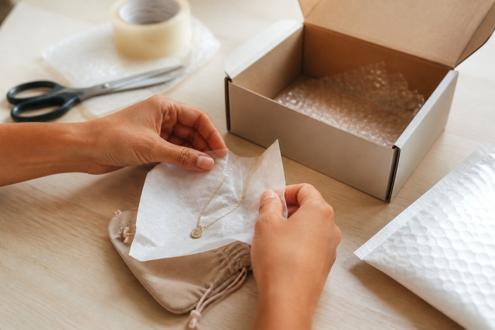 Hands carefully wrapping a delicate gold necklace with tissue paper and placing it into a small protective pouch, with a padded shipping box and bubble wrap visible on a clean work surface