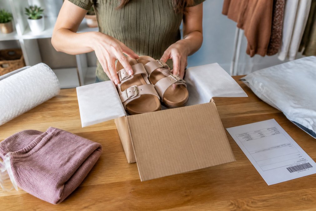 A woman folding a pair of women's fashion sandals with adjustable buckles and placing them into a plain cardboard shipping box on a wooden table, with a printed return label and packing slip visible nearby