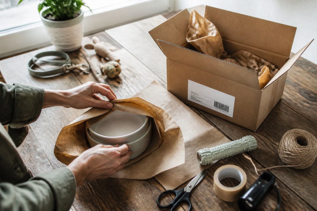 Hands carefully wrapping pet products in recycled kraft paper and packing materials, with a cardboard box and shipping label visible on a clean wooden workspace, natural window lighting