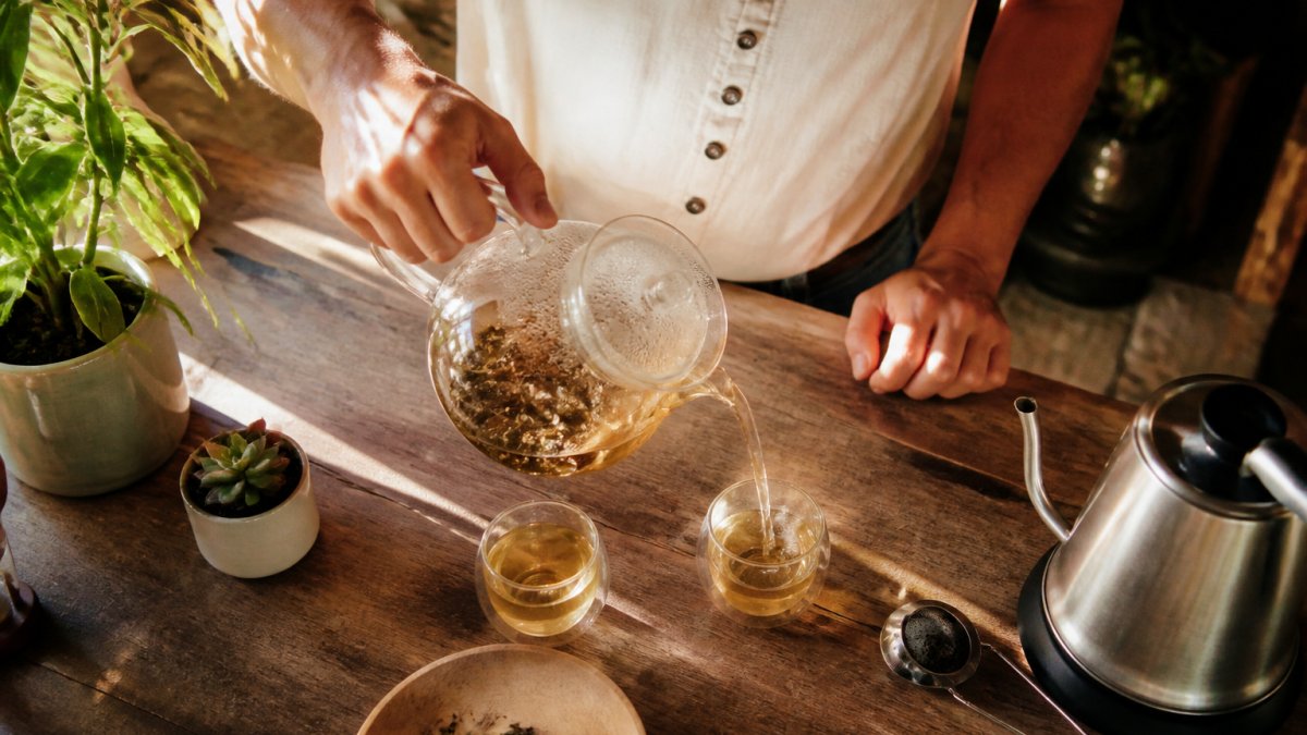 Glass teapot with spent loose leaf tea on a wooden kitchen counter with a cup of tea being poured nearby.