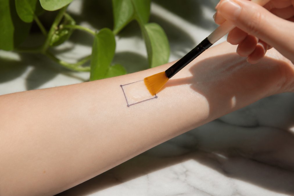 Close-up of person's forearm showing a small marked patch test area with chemical peel solution applied using a brush applicator on clean, pale skin in natural daylight