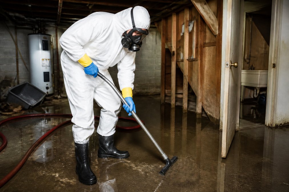 Professional wearing complete protective equipment including fitted respirator mask, double gloves, protective suit covering entire body, and rubber boots in contaminated basement environment with visible standing water