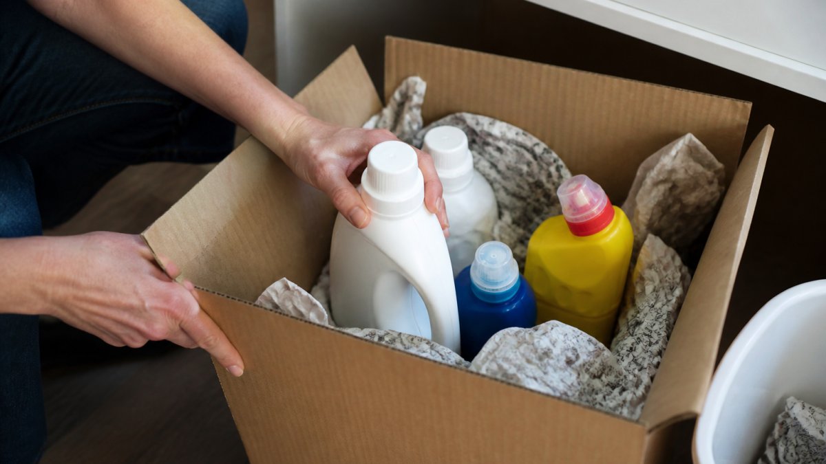 Close-up of hands carefully packing cleaning product bottles in a sturdy cardboard box with protective padding and newspaper, showing proper containment before transport to a collection site