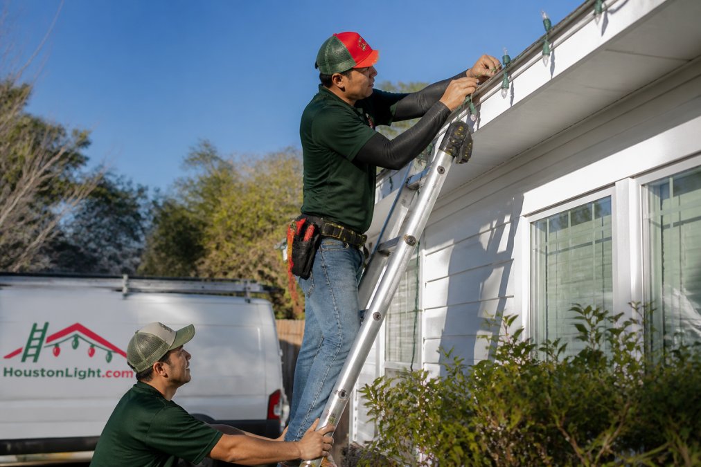 A person on an extension ladder secured against a home's roofline, attaching LED light strands to the eaves using plastic shingle clips, with a spotter standing at the base of the ladder on a clear day