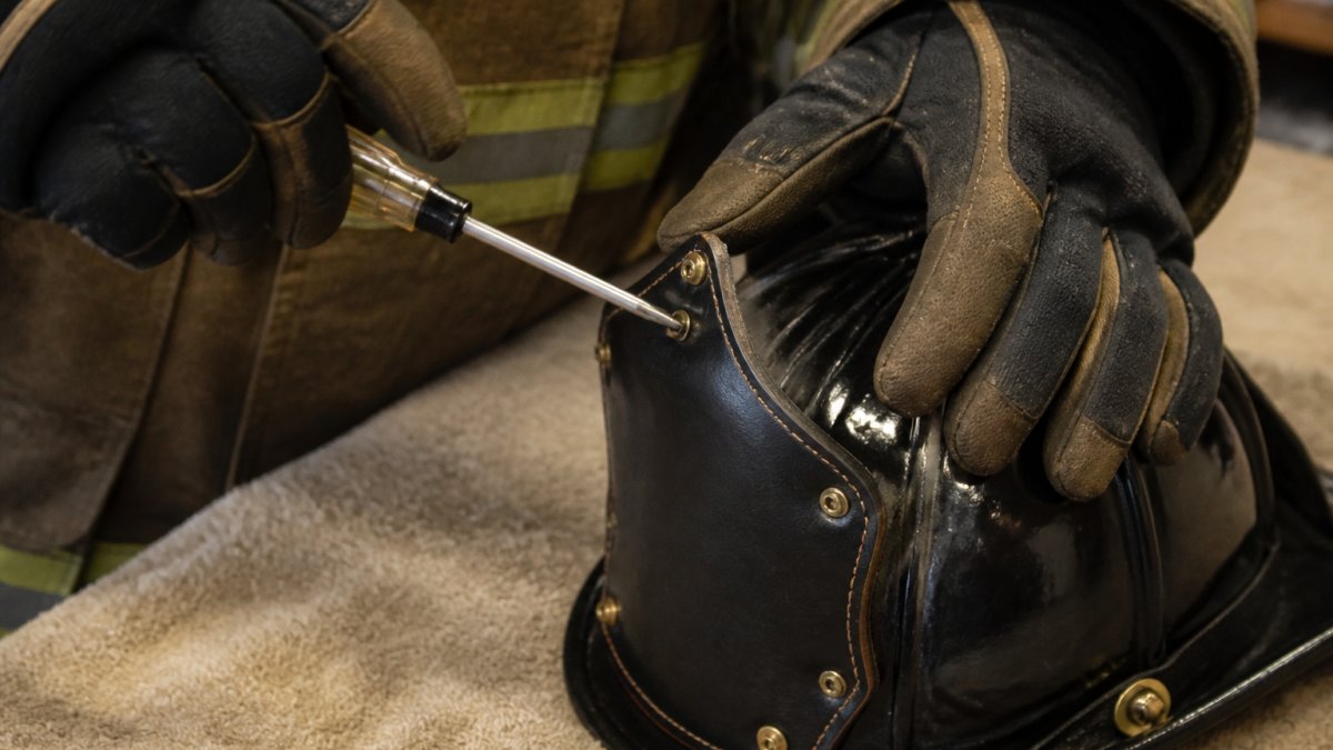 Close-up of a firefighter's gloved hands using a small hand screwdriver to carefully loosen brass screws on a leather fire helmet front holder, soft workshop lighting, towel-covered bench