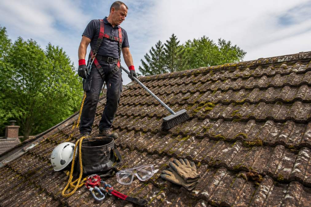 A homeowner wearing a safety harness and non-slip shoes standing on a tiled roof, holding a soft-bristle brush, with safety equipment visible nearby