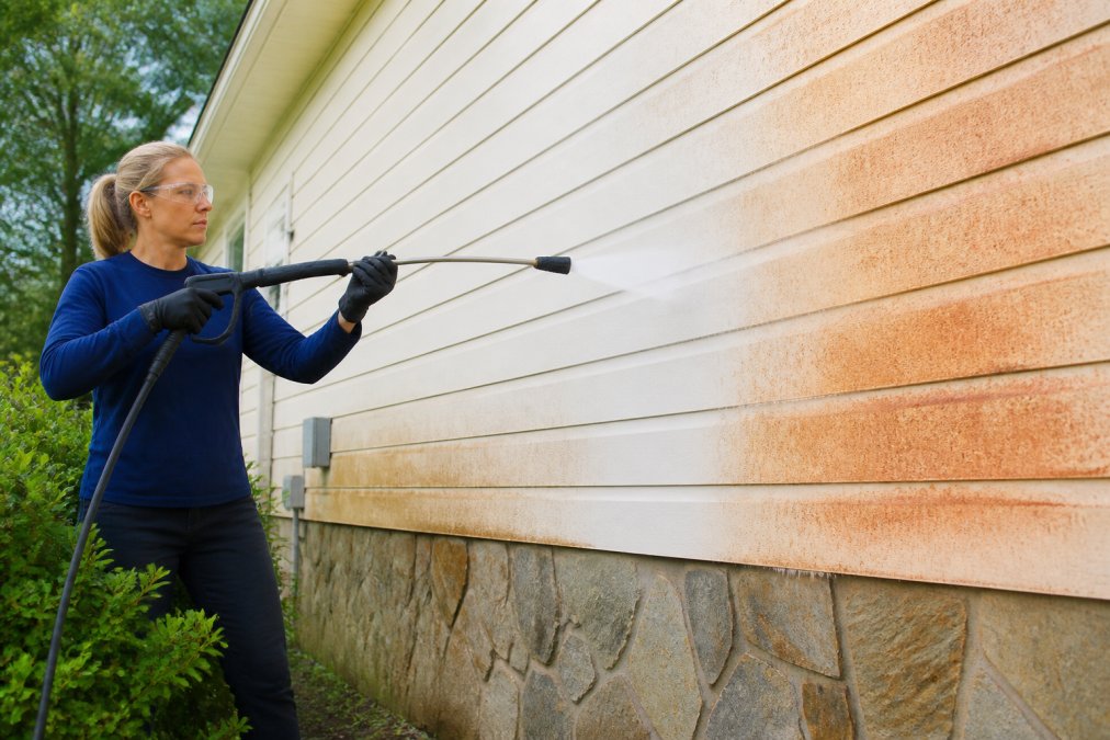 Homeowner carefully spraying siding with a soft wash nozzle, showing a gentle water stream hitting a clay-stained exterior wall in natural daylight, protective equipment visible