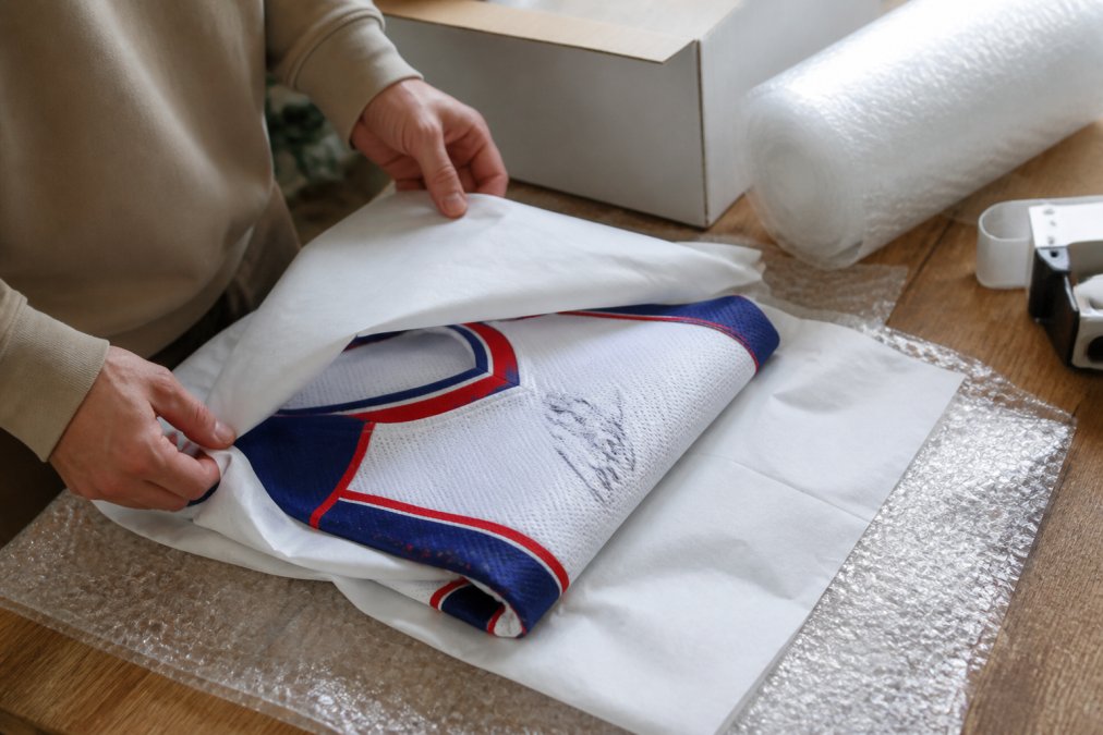 Hands carefully wrapping a vintage signed hockey jersey with acid-free tissue paper and bubble wrap on a wooden work surface with natural lighting