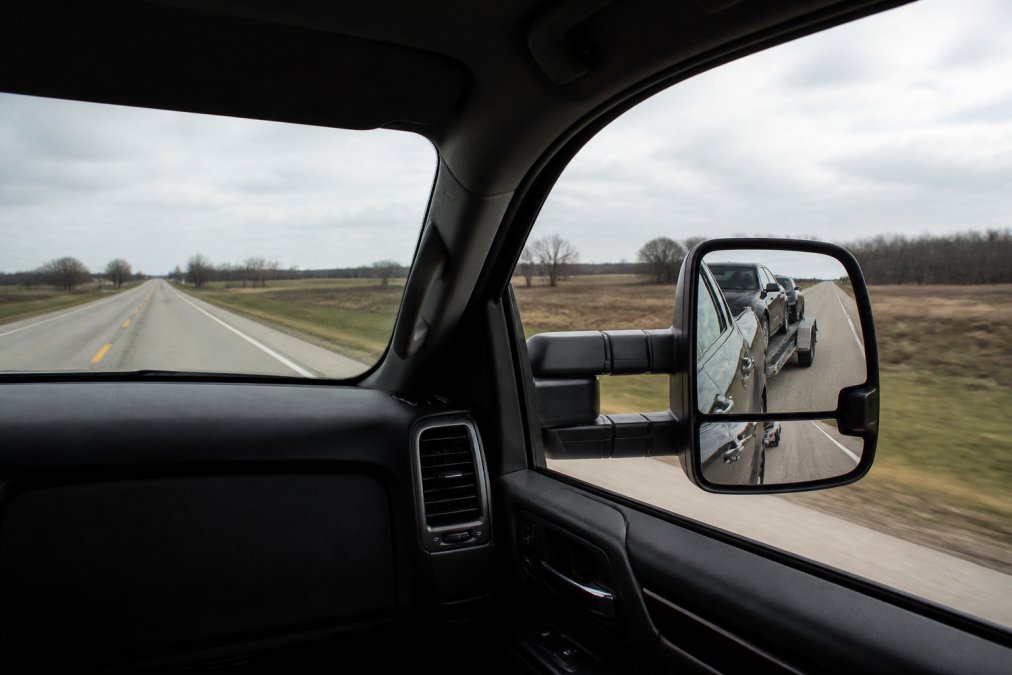 View from inside a pickup truck cab towing a loaded open car hauler on a two-lane highway, wide towing mirrors mounted on both doors showing the trailer tracking straight behind, overcast daylight