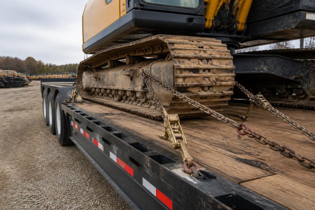 Heavy-duty chains and ratchet straps securing an excavator to a lowboy trailer at a construction yard, binders tight and chains crossing over the tracks at proper tie-down points
