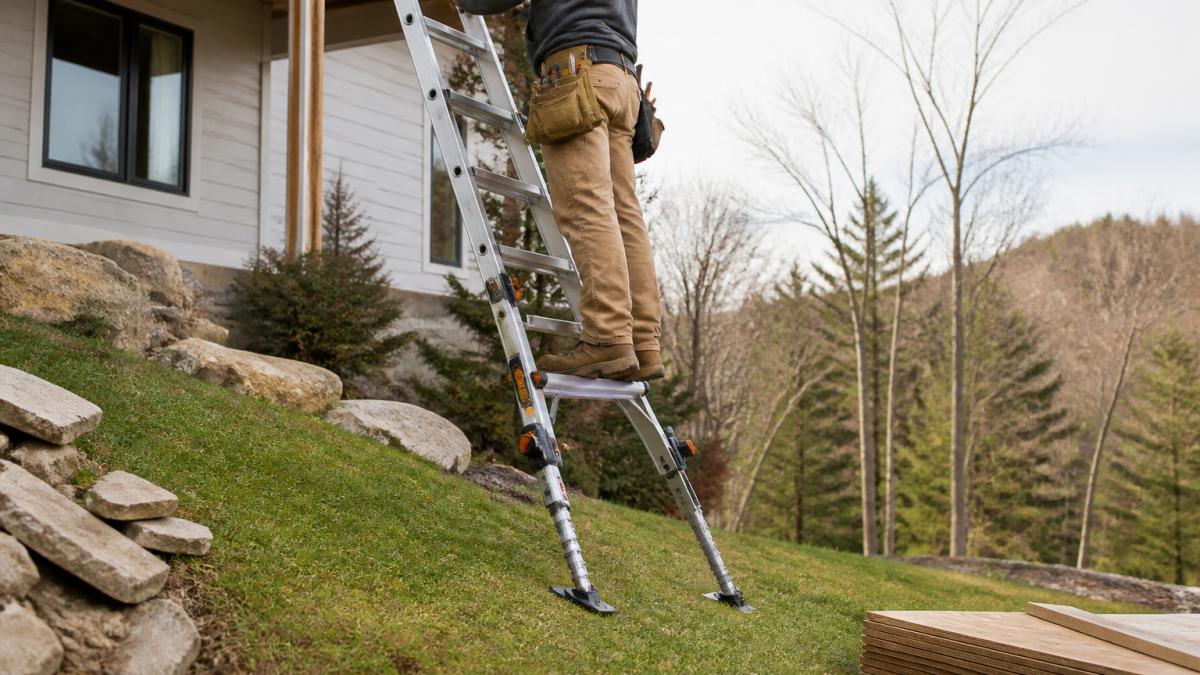A contractor in work clothes and safety boots using a multi-position ladder with adjustable leveling legs on a grassy slope, the ladder's feet clearly planted on firm ground