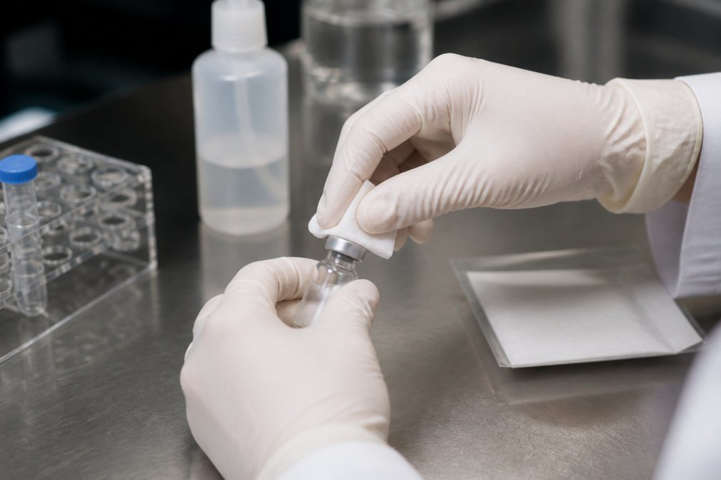 Researcher's gloved hands using a sterile alcohol pad to swab a peptide vial rubber stopper with firm circular motions on a clean laboratory work surface under bright task lighting