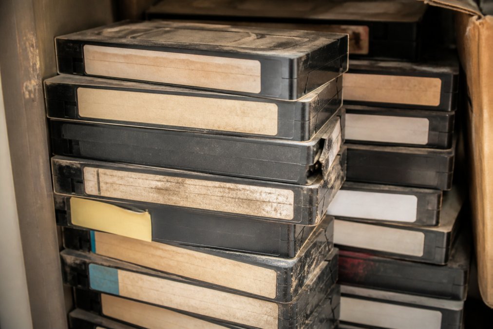 Stack of aged VHS tapes showing visible wear, dust, and discolouration on spines, stored in poor conditions with faded labels