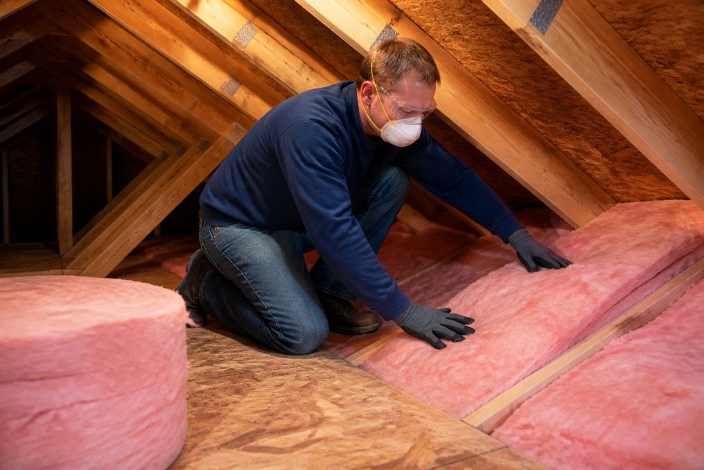 A technician in protective work gear and gloves pressing insulation batts into the joist cavities of a residential attic, with rolls of pink fiberglass insulation visible on the plywood floor nearby under warm work lighting
