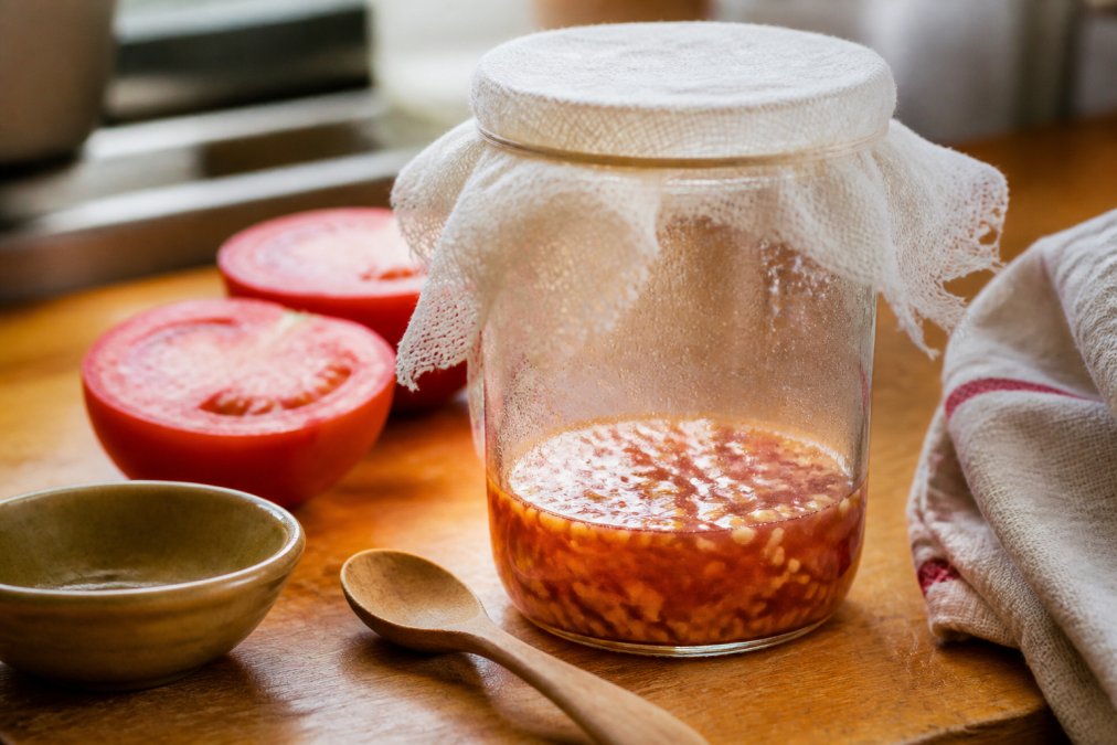 Close-up of a mason jar filled with tomato seeds and pulp in water on a wooden kitchen counter, with cheesecloth loosely draped over the top and a handwritten label nearby