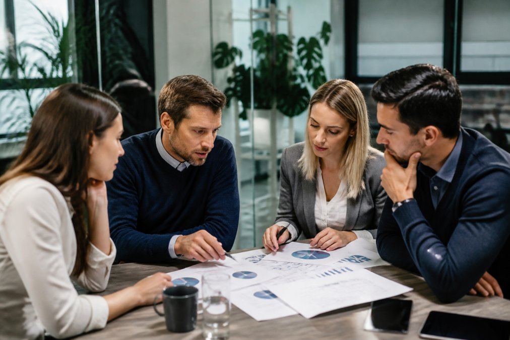 Marketing team reviewing analytics dashboard on multiple monitors in modern office environment, with team members pointing at KPI metrics and revenue charts on screens