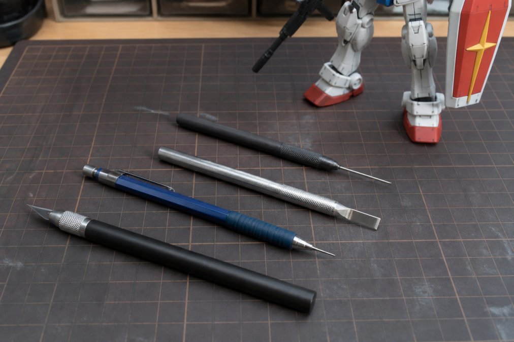 Flat lay of various scribing tools including hobby knife, mechanical pencil, chisel-style scriber, and needle-style scriber arranged on a clean work surface with scale model in background
