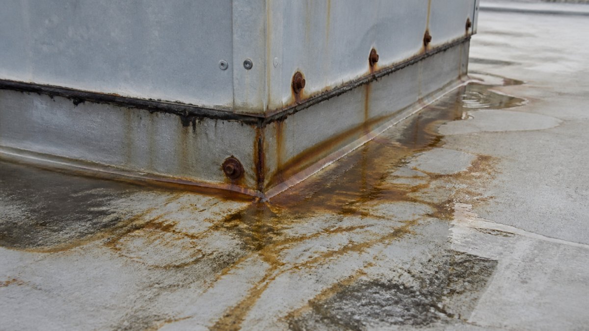 Close-up of a roof hatch with visible water stains and corrosion around the frame and fasteners, showing active leaking and deteriorated seals