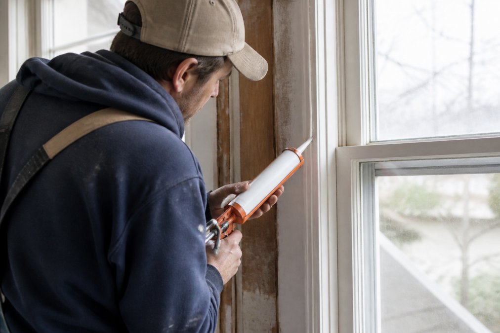 Contractor using a caulking gun to apply sealant around a window frame in an older home, showing proper 45-degree angle technique and smooth, continuous bead application