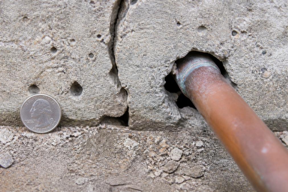 Close-up of a concrete foundation showing a visible crack and small gaps around a copper pipe penetration, with a quarter-inch reference object for scale