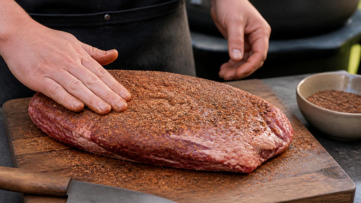 Close-up of hands patting a dark spice rub onto a raw brisket flat on a wooden board, with a small bowl of rub and a knife nearby