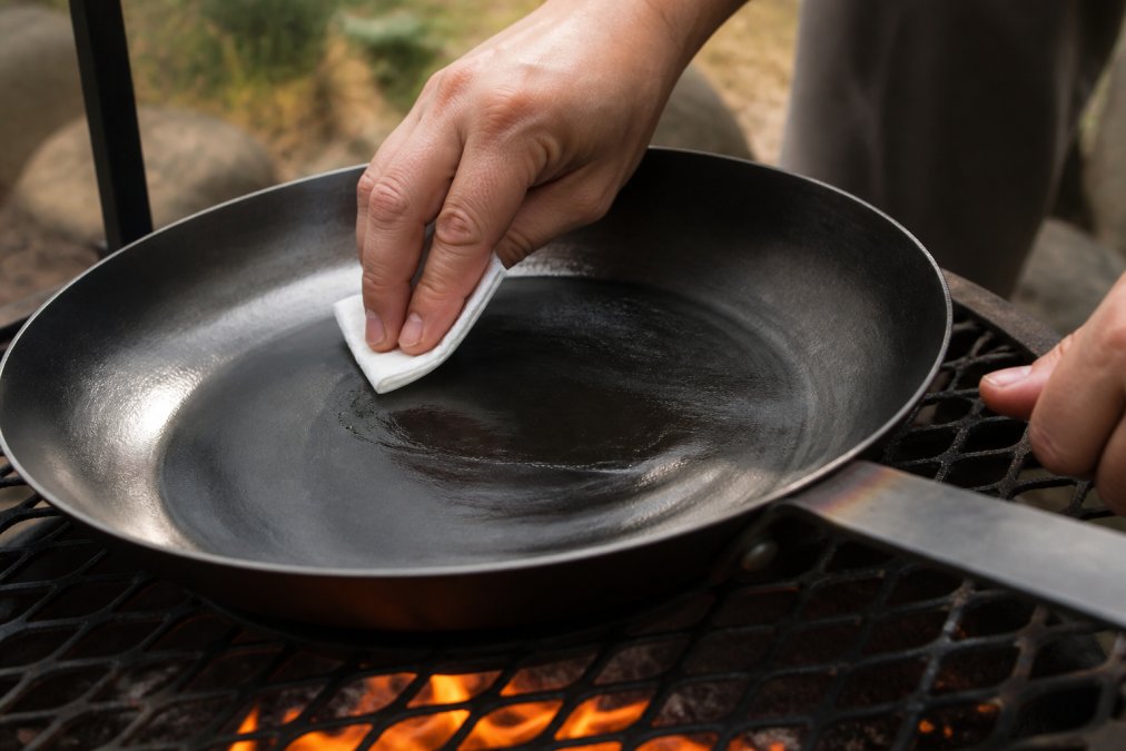 Hands buffing a carbon steel pan with a white paper towel over a stovetop burner with orange flame visible, showing the thin layer application technique in close-up