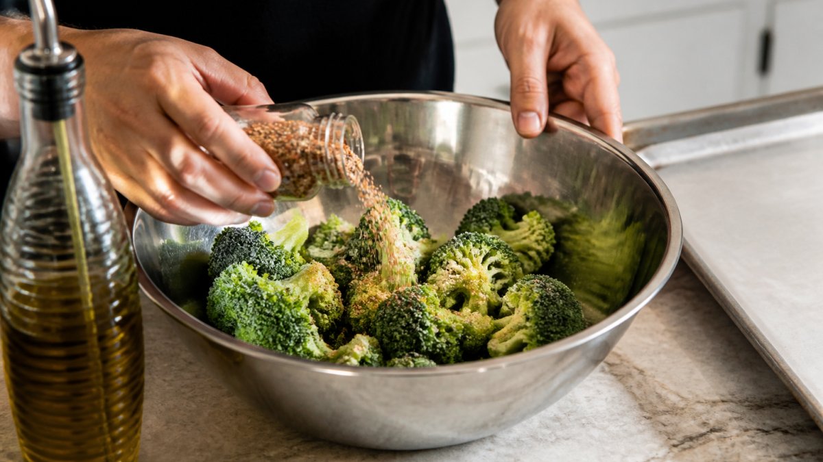 Hands tossing frozen broccoli florets with olive oil and spices in a large mixing bowl on a kitchen counter