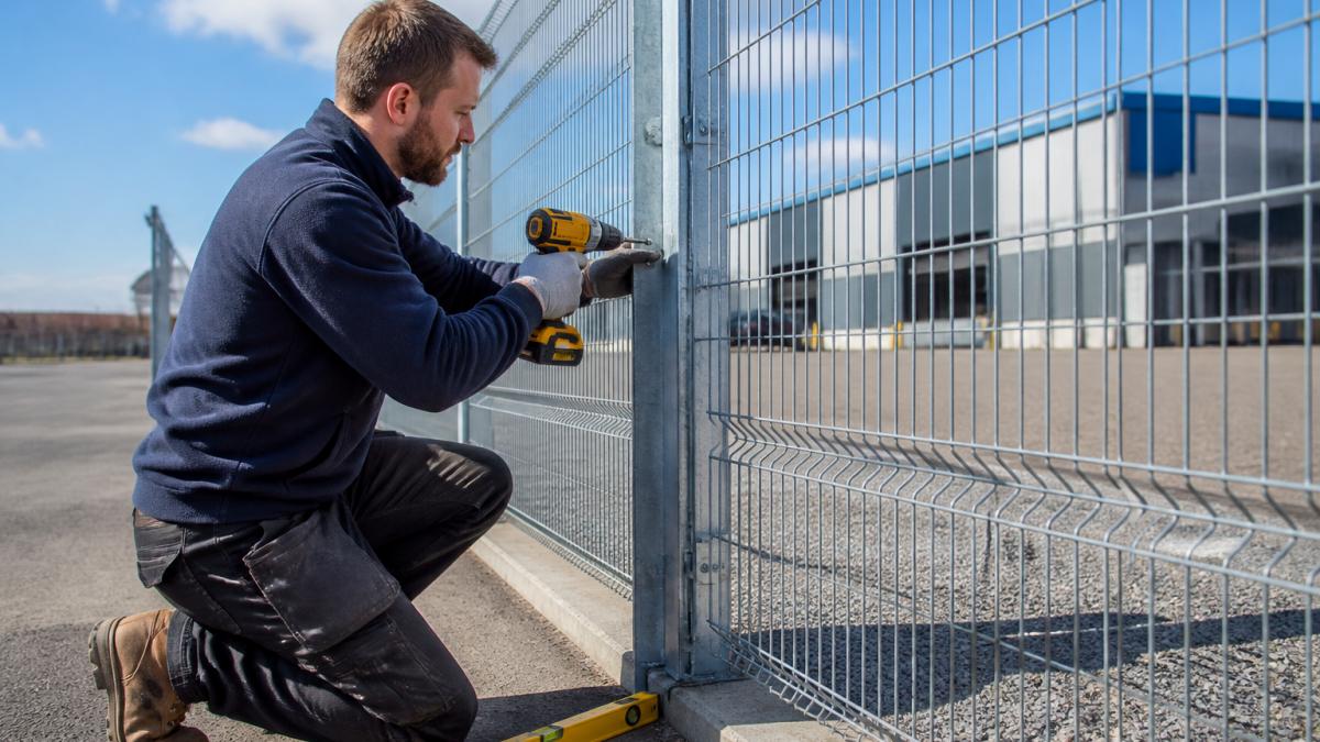 A professional fence installer in work clothes and safety gloves securing a galvanized steel anti-climb mesh panel to a metal post using a drill, with a level and tape measure visible on the ground, set against a commercial warehouse perimeter on a clear day