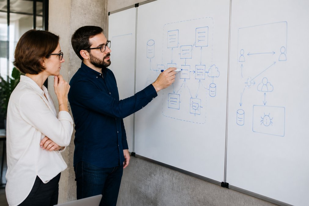 Security architect and AI engineer reviewing agent architecture on whiteboard in modern office, with diagrams of system components and threat vectors visible on the board