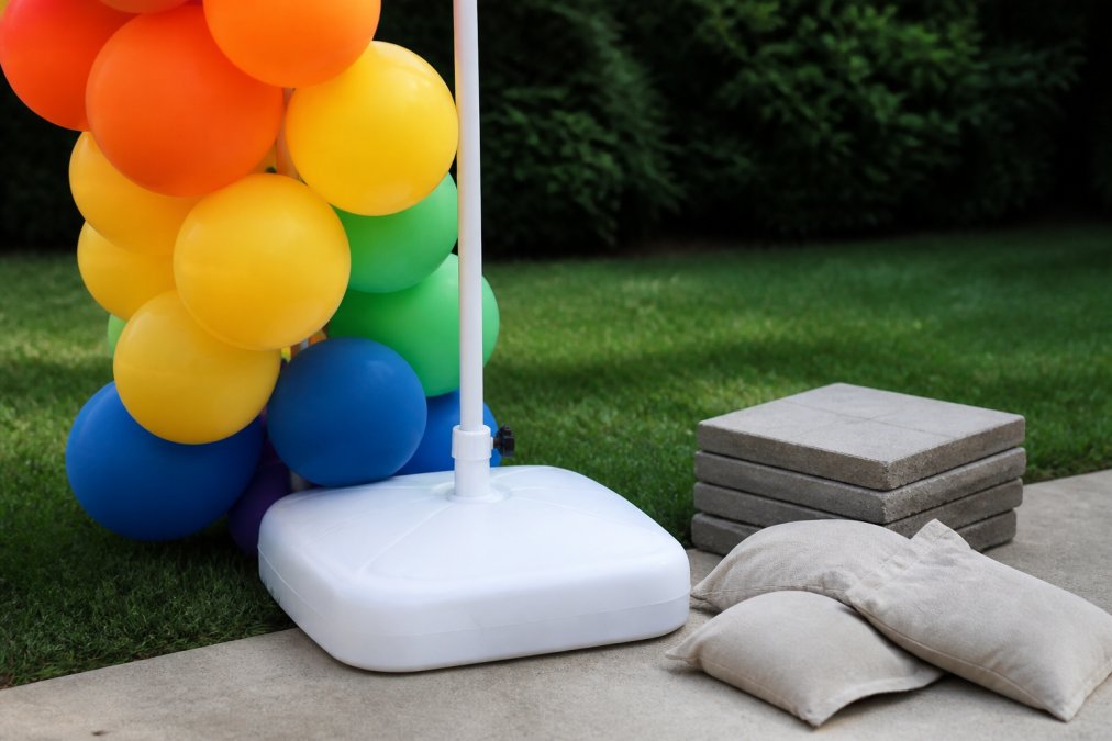 Water-filled base plate securing outdoor balloon decor on a lawn with sandbags and pavers nearby