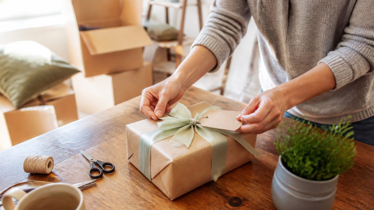 A person arranging a wrapped housewarming gift with a handwritten note and ribbon on a wooden table, surrounded by moving boxes and a small plant