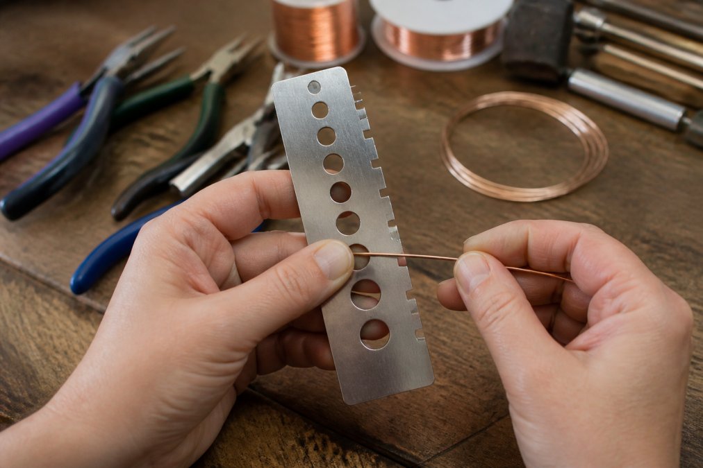 Close-up of a crafter's hands using a metal wire gauge tool to measure a piece of copper wire on a wooden workbench, with jewelry pliers and wire spools visible in the background