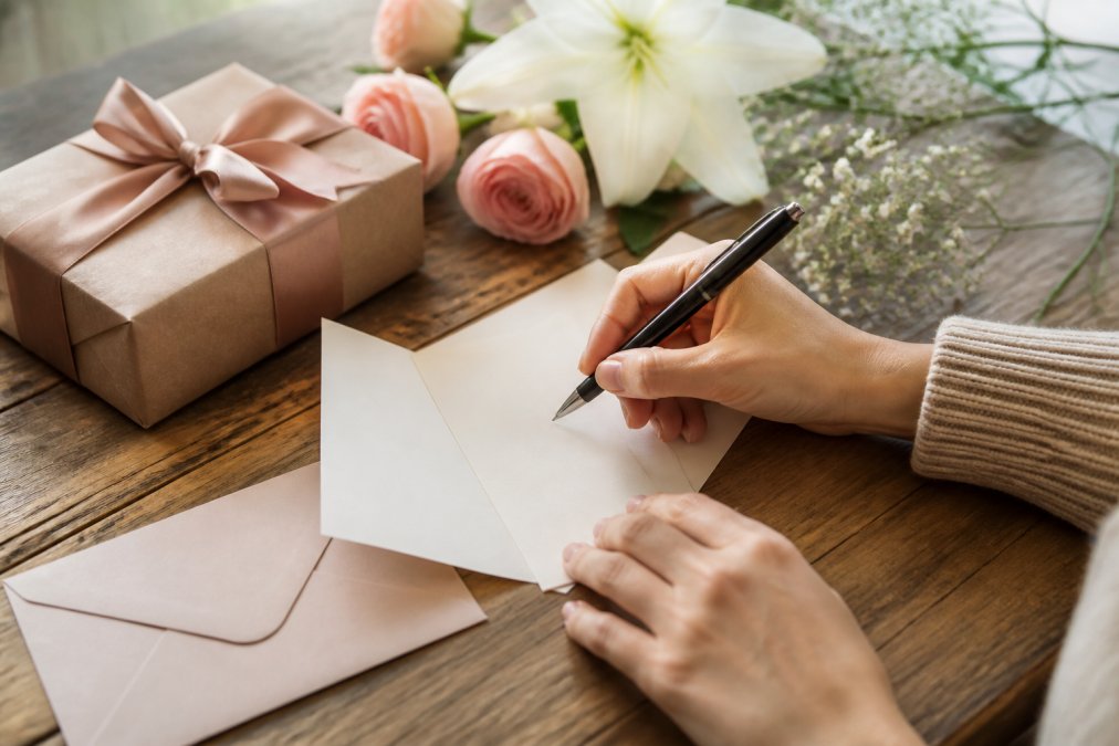 Hands writing a handwritten note with a pen on elegant stationery, with a wrapped gift and flowers visible in the background, soft natural lighting