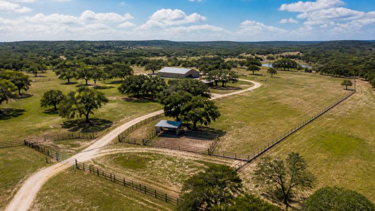 Wide aerial view of a rural ranch property with well-maintained fencing, open pasture, scattered trees, and clear property boundaries visible under bright Texas sky