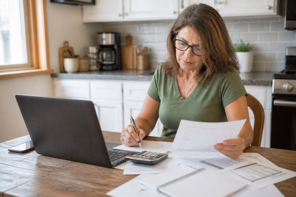 Homeowner reviewing documents and taking notes at a kitchen table with a laptop and phone nearby, natural lighting from window, papers and calculator visible