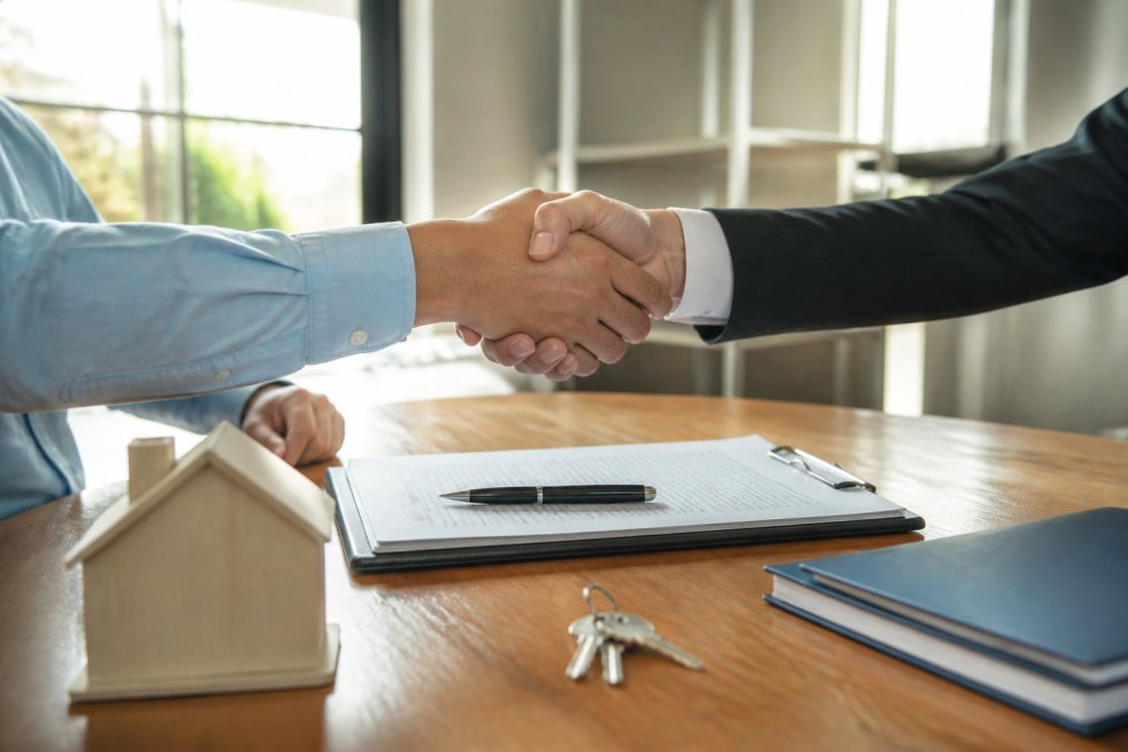 Two people shaking hands across a desk with title document and pen visible, professional office setting with natural lighting, documents and folder on desk