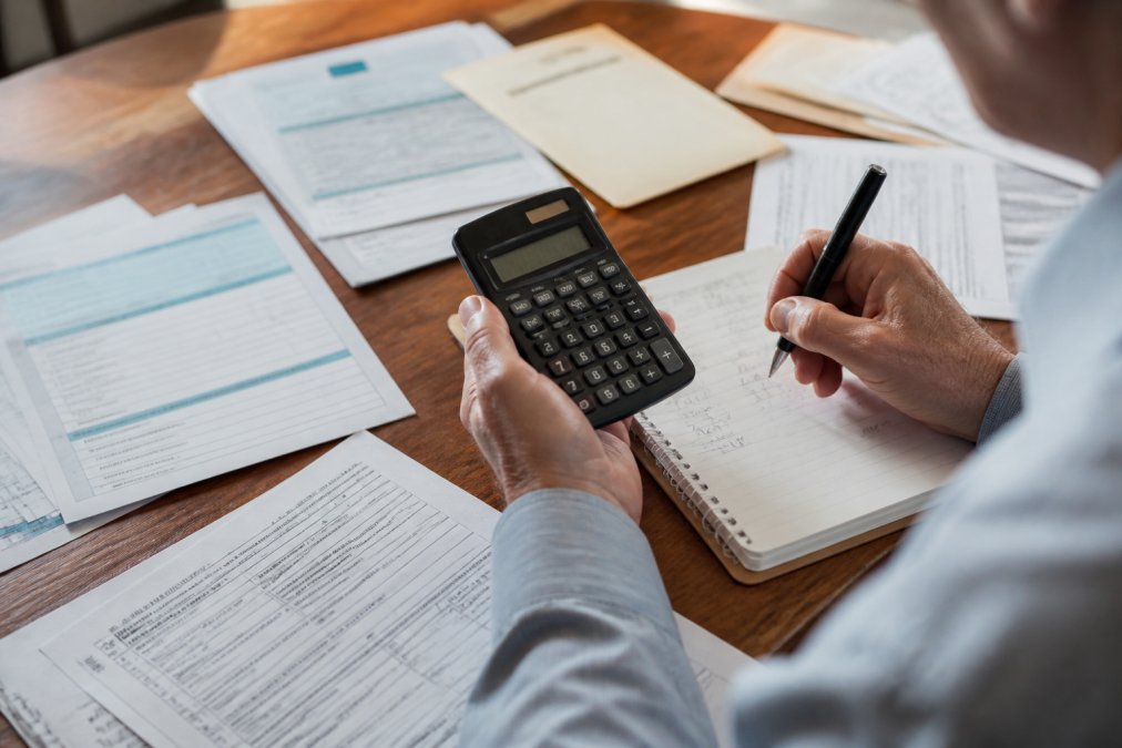 Close-up of hands holding a calculator and reviewing retirement withdrawal calculations on a notepad, with tax forms, account statements, and documents spread across a desk under natural lighting