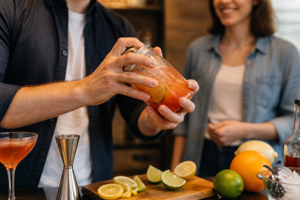 Close-up of hands tightly holding a glass jam jar filled with ice and a colourful cocktail mixture, being shaken over a kitchen counter with citrus slices nearby