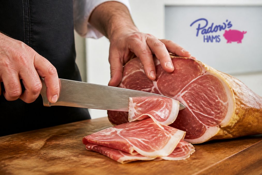 Close-up of a sharp knife thinly slicing a salt-cured country ham on a wooden carving board, showing the proper angle and technique for thin slices