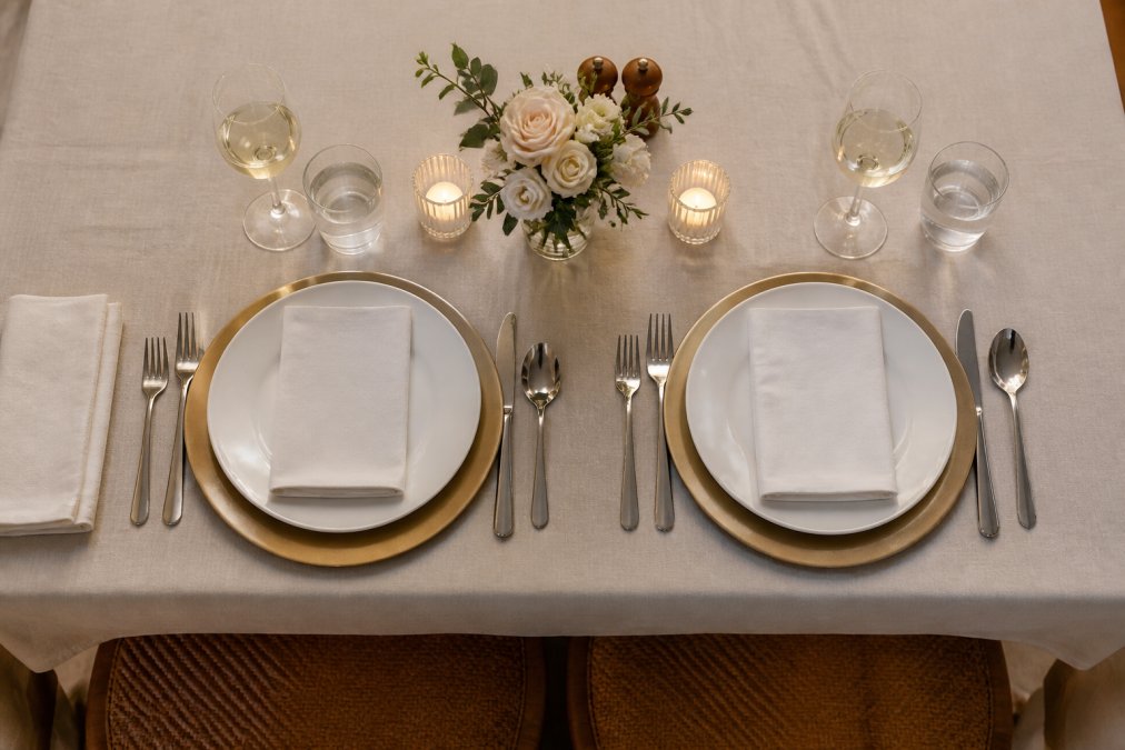 Close-up overhead view of an elegantly set place setting for two with properly positioned cutlery on either side of a charger plate, white wine and water glasses positioned above the knife, and a folded linen napkin placed to the left of the forks on a neutral cream tablecloth
