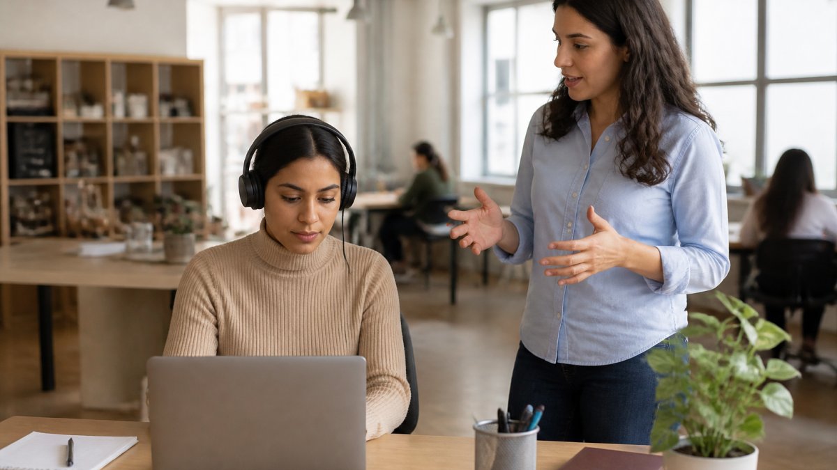 A woman at her desk wearing headphones, focused on her laptop, while a coworker stands nearby talking in a bright open-plan office