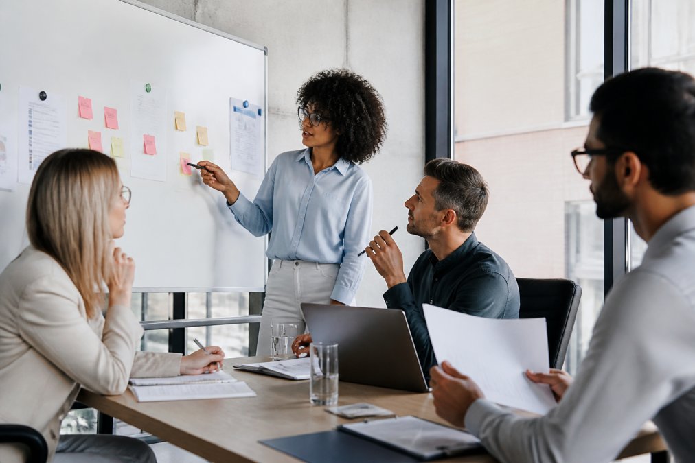 Professional team in modern conference room reviewing strategic business objectives on whiteboard, with team members pointing at goals and discussing event planning approach